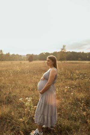 Expectant mother stands in profile amid golden sunset field wearing flowing dress.
