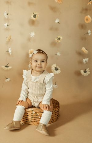 Baby in white outfit sits on wicker basket with floral backdrop.