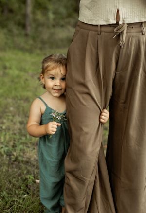Small child in teal overalls peeks out from behind adult wearing brown pants outdoors.