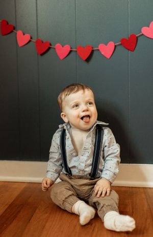Baby in suspenders sitting against wall with red heart garland decoration above.