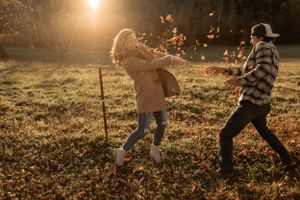 A couple playing in fallen leaves during sunset in a rural setting.