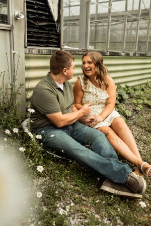 A romantic couple sitting together near a greenhouse surrounded by small white flowers.