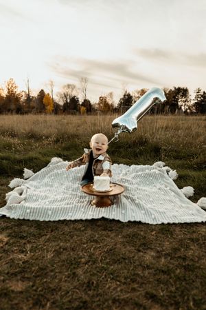 A baby sits on a white blanket in a rustic field during golden hour with blue birthday decorations.