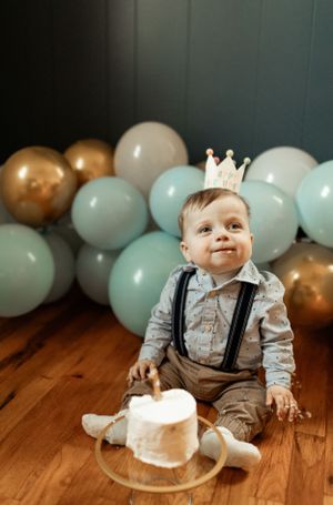 Baby's first birthday setup with crown, balloons, and smash cake on hardwood floor.