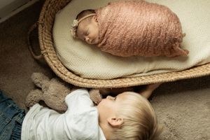 Two small children looking at sleeping newborn in woven basket on cream colored cushion.