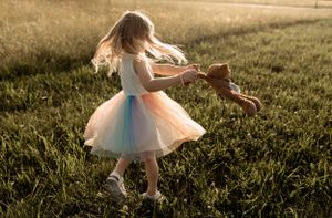 Child dancing in tutu dress with teddy bear during sunset in field.