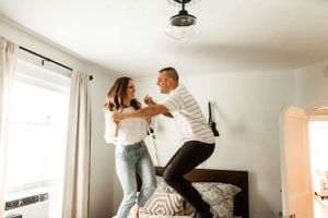 A playful couple jumps on a bed together in a sunny bedroom.