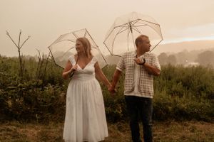 A couple holds clear umbrellas while standing in a misty field at sunset.