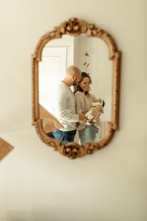 A reflection in an ornate gold mirror shows a tender family moment in a bright room.