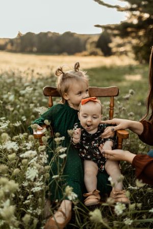 Two children interact while sitting in a wooden chair among wildflowers at sunset.
