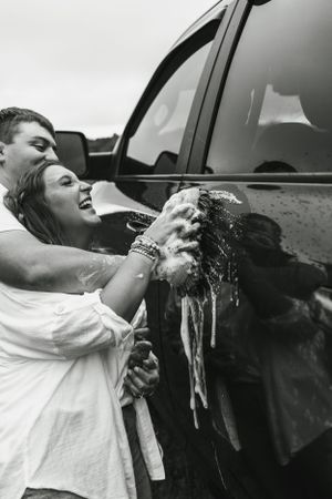 A playful couple washing a black truck together in black and white.