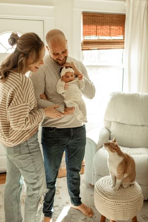 A couple holds their newborn baby in a sunny room while their orange cat watches from a knit ottoman.