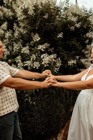 A couple holds hands in front of blooming white flowers during an engagement shoot.