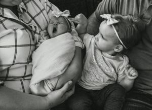 Black and white photo of a toddler looking adoringly at a swaddled newborn baby in a tender moment.