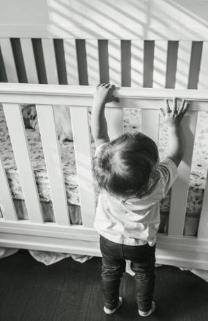 Toddler reaching up to white crib rails with sunlight streaming through window blinds.