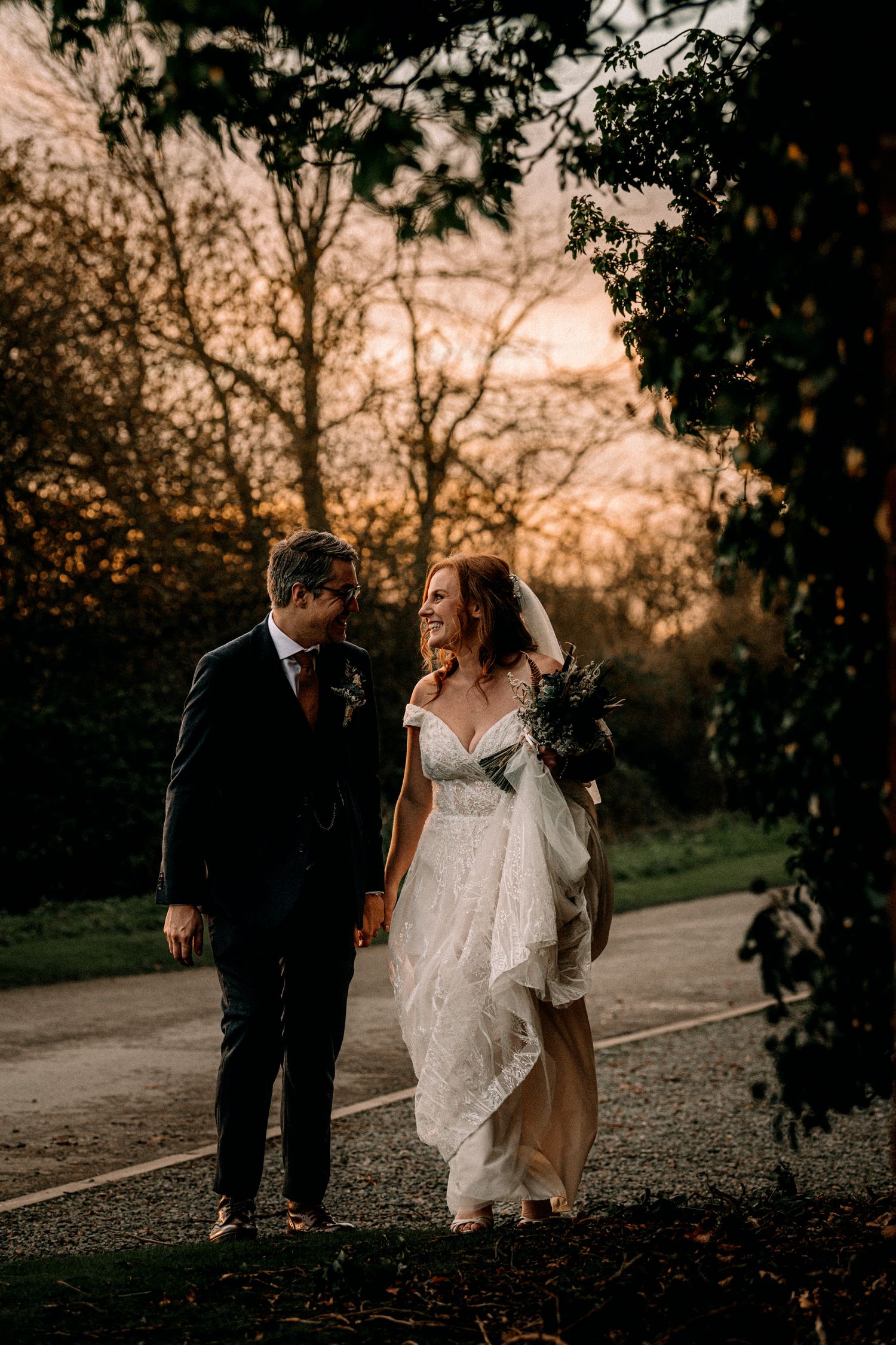 A couple in wedding attire walk together on a path during sunset as golden light filters through autumn trees.