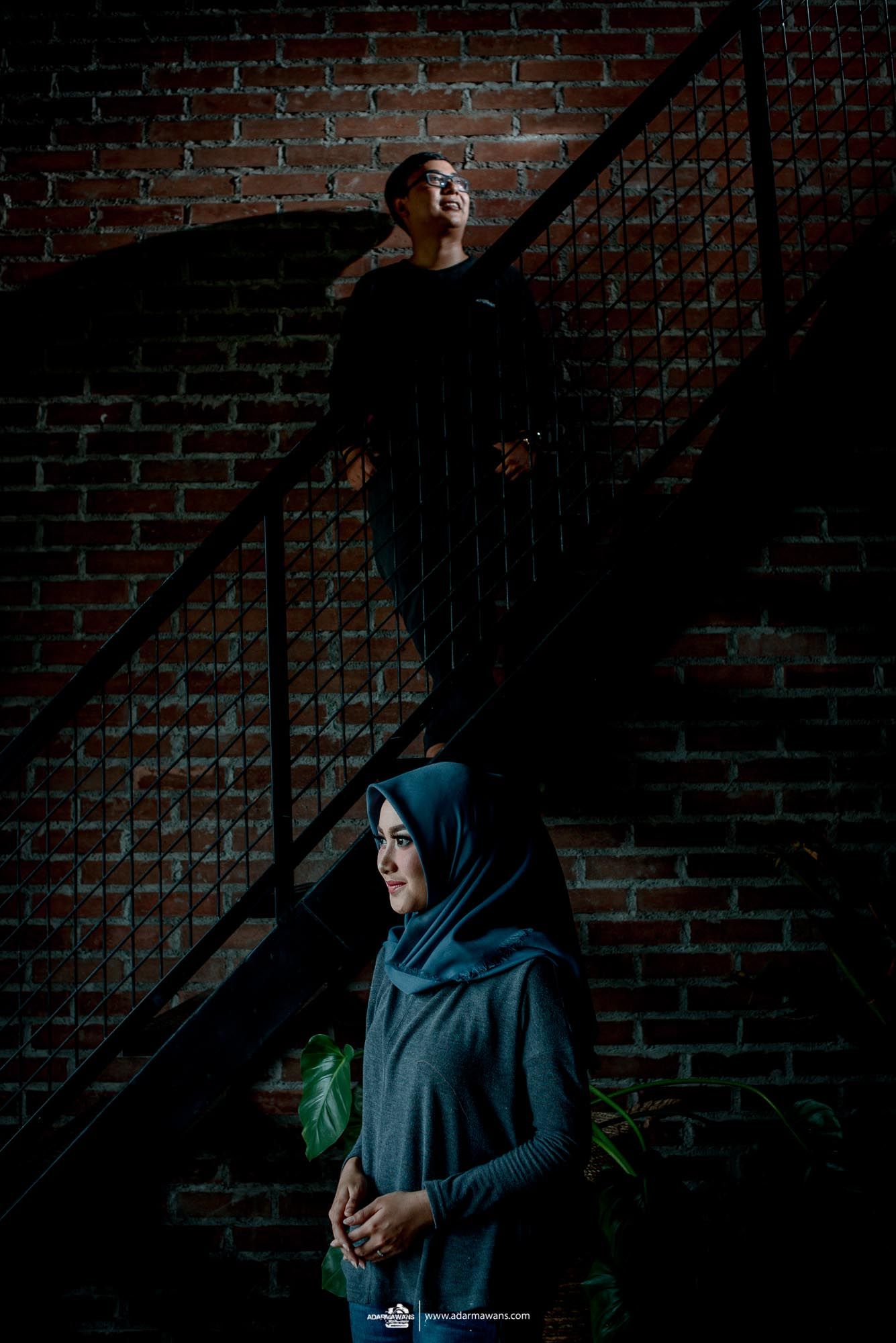 Two people pose dramatically on a staircase against a brick wall in moody lighting.