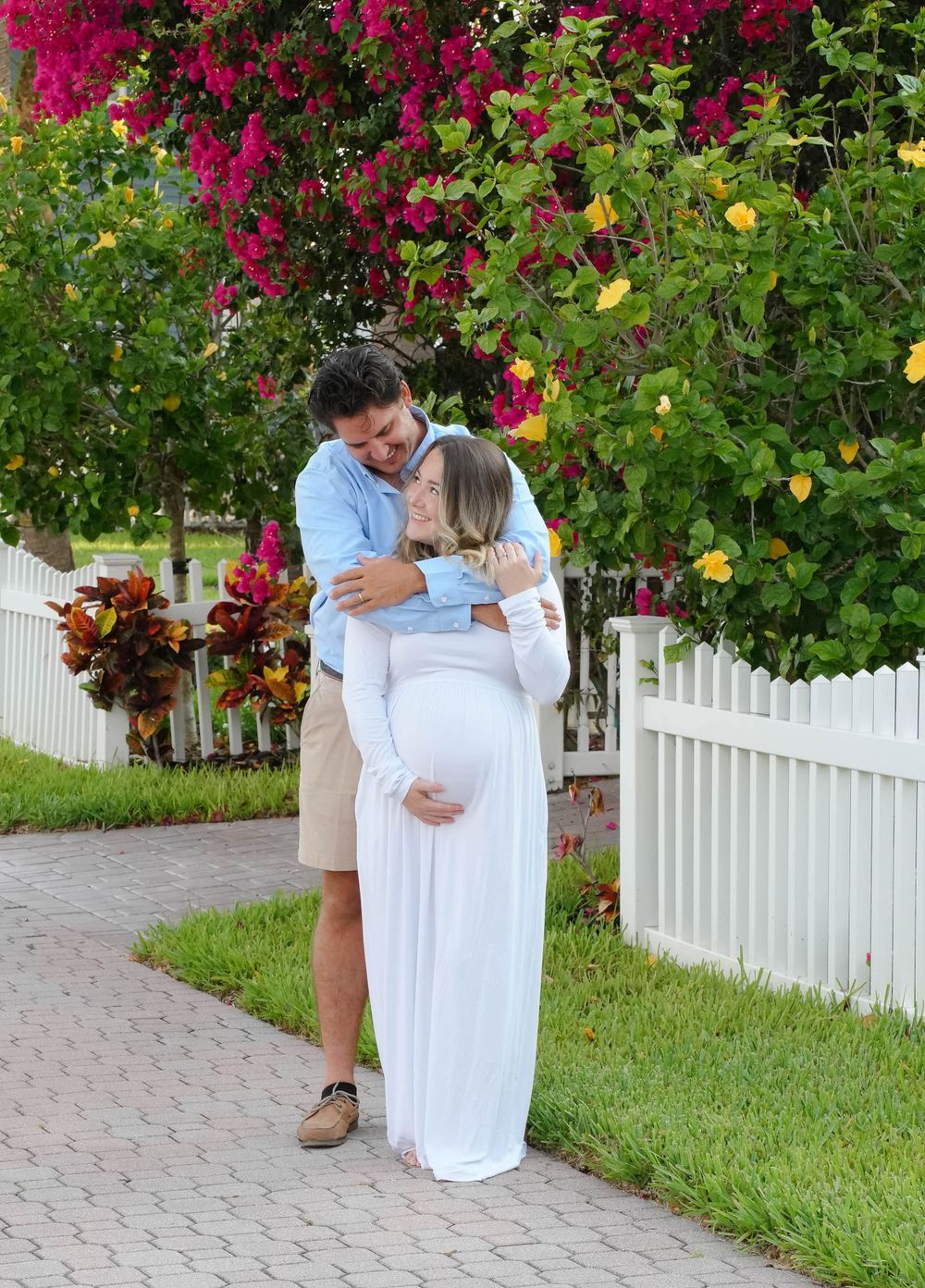 A tender moment with expecting parents along a picturesque garden path with white picket fence and vibrant pink bougainvillea flowers.