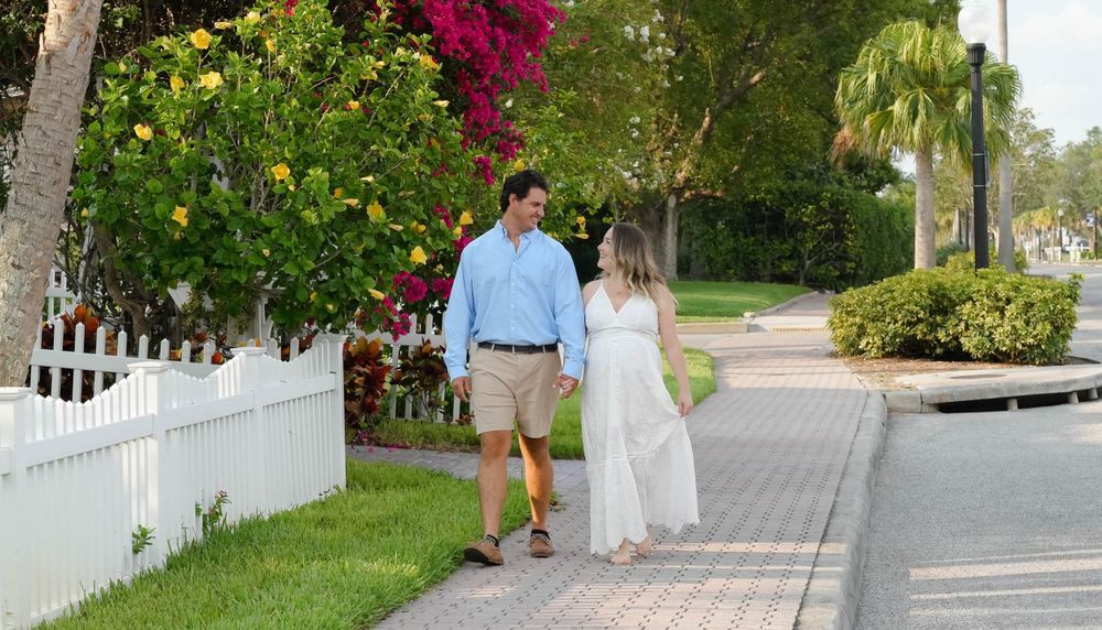 Expecting couple walking along a residential sidewalk with a white picket fence and vibrant bougainvillea flowers in Florida.