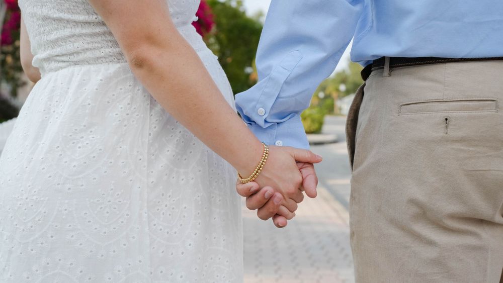 Expecting couple holds hands with coastal homes in the background.