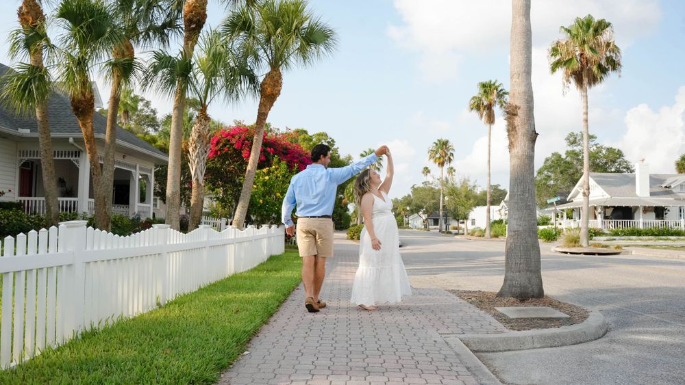 Expecting couple dances on a palm-lined sidewalk with white picket fences and coastal homes in the background.