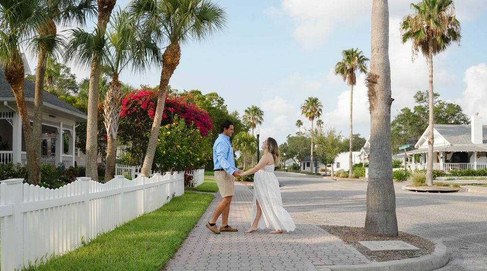 An expecting couple walks hand in hand down a palm tree-lined street with white picket fences in a tropical neighborhood.
