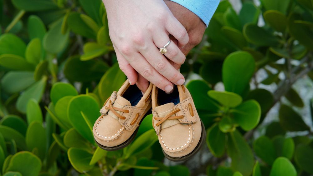 Soon to be parents hold hands with tiny beige leather baby moccasins against a green leafy background.