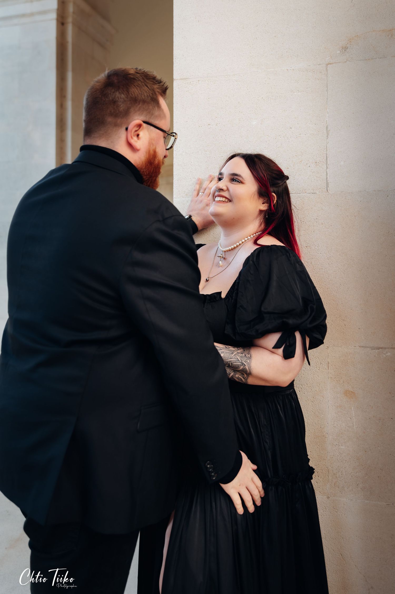 Couple in matching black outfits sharing a tender moment against stone column.