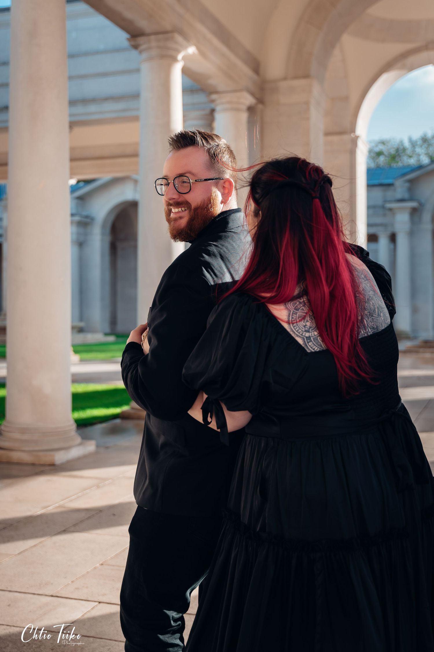 Smiling bearded man in black suit embracing red-haired woman at columned monument.