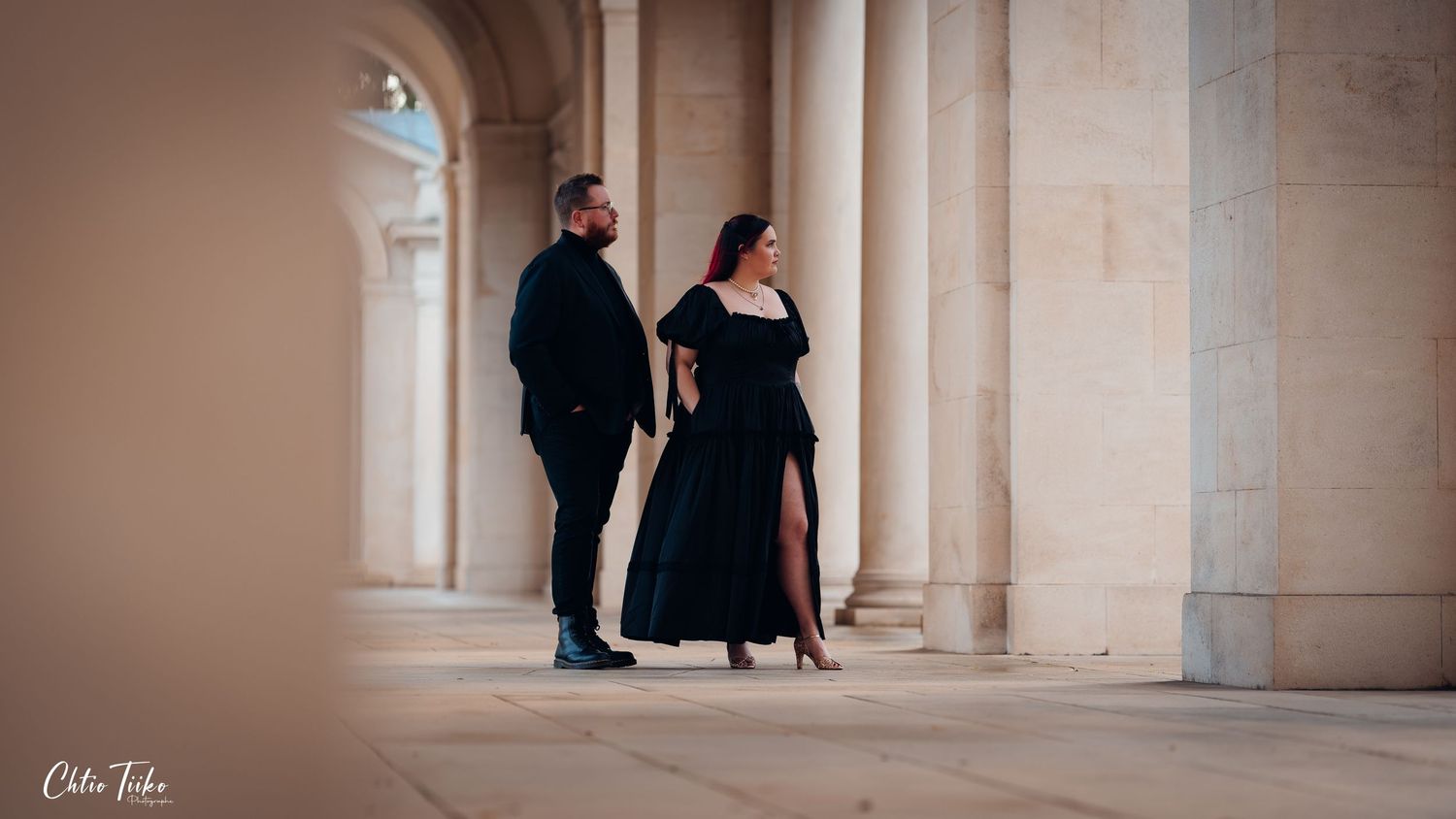 Elegant couple in black formal wear posing between grand stone columns.