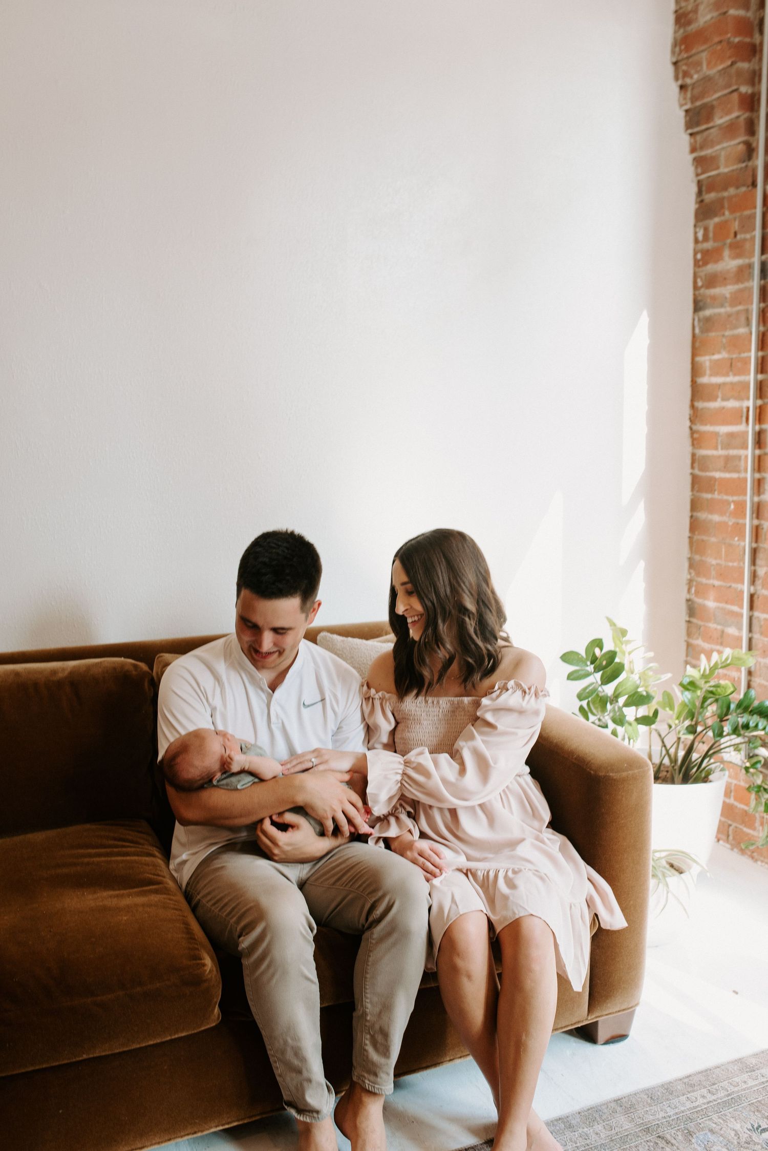A couple sits together on a brown couch against a brick wall in a bright room with natural lighting.