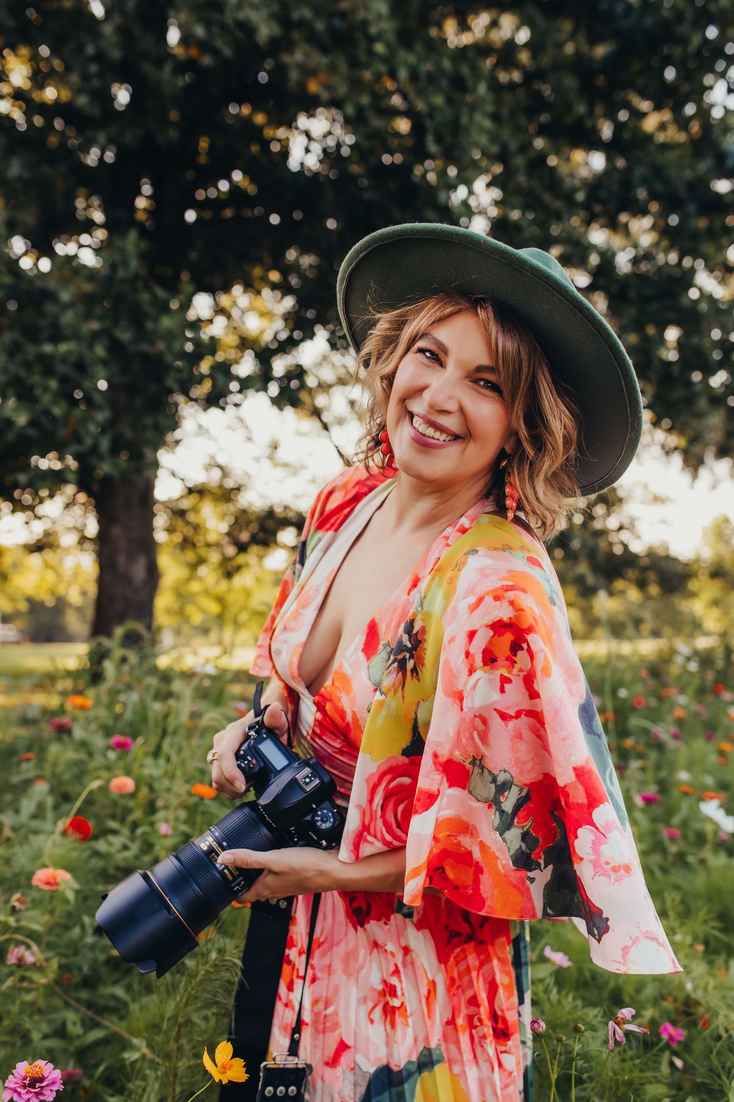 A photographer in a floral dress and hat stands among blooming flowers at golden hour.