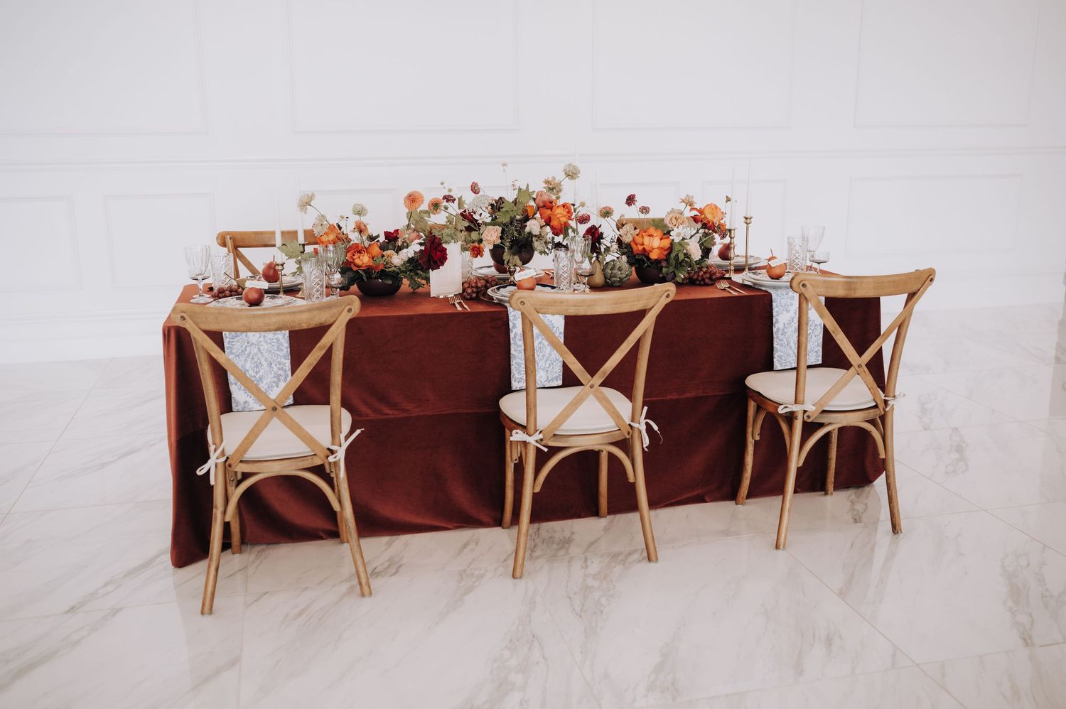 Rustic wedding table setup with wooden crossback chairs and terracotta tablecloth decorated with floral arrangements.