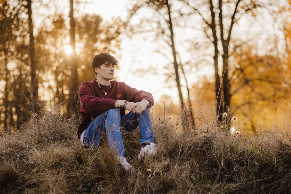 Silhouette of person sitting in tall grass during golden sunset with autumn trees in background.