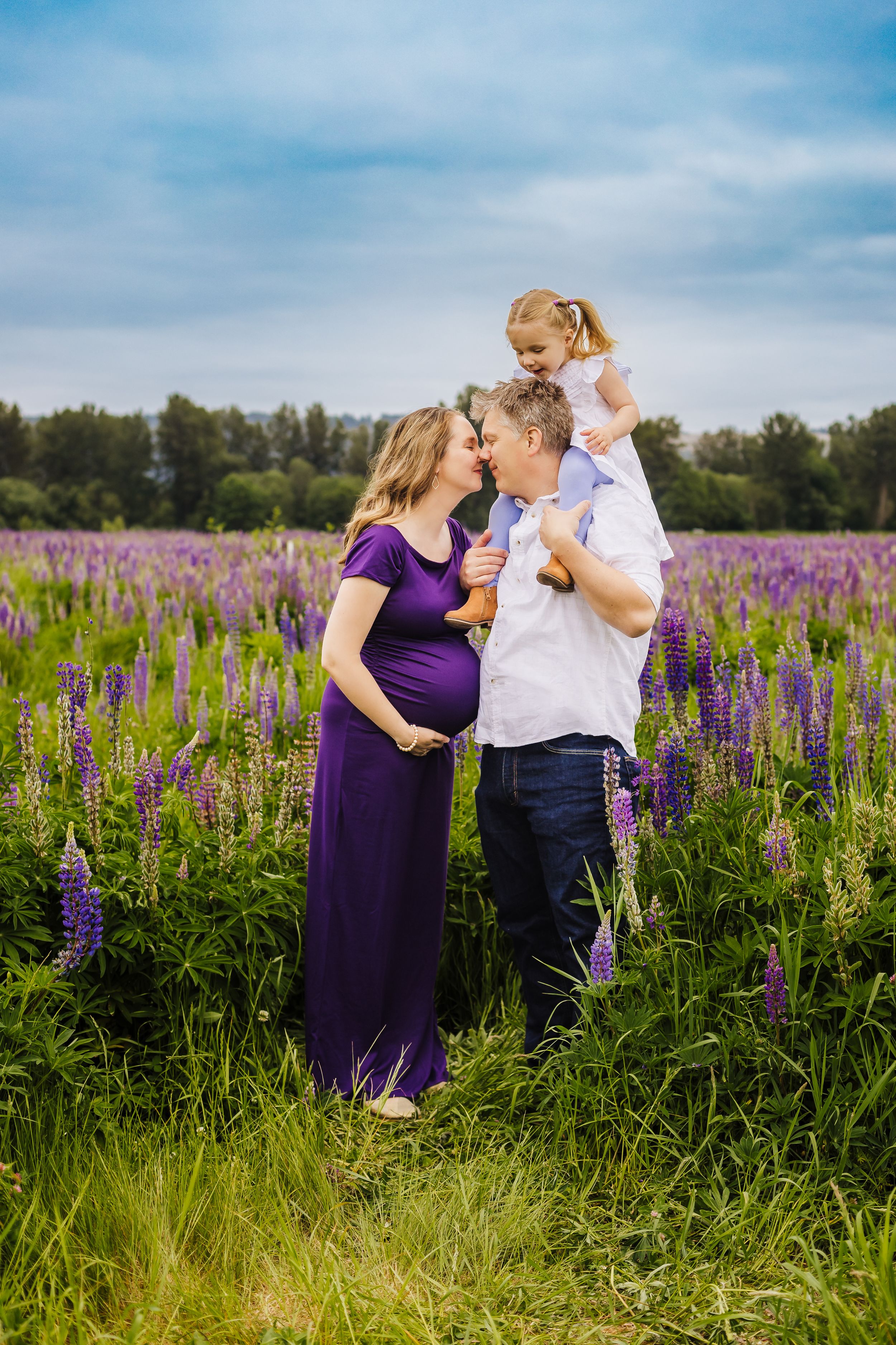 Family shares tender moment in purple lupine field with expectant mother in purple dress.