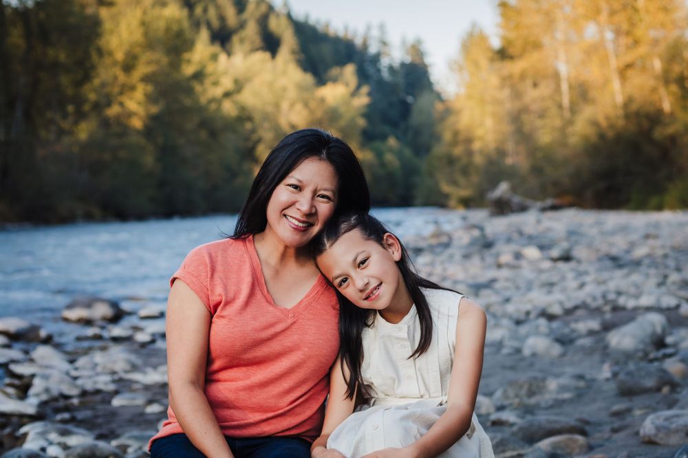 Mother and daughter smiling together by rocky riverbank with autumn trees in background.