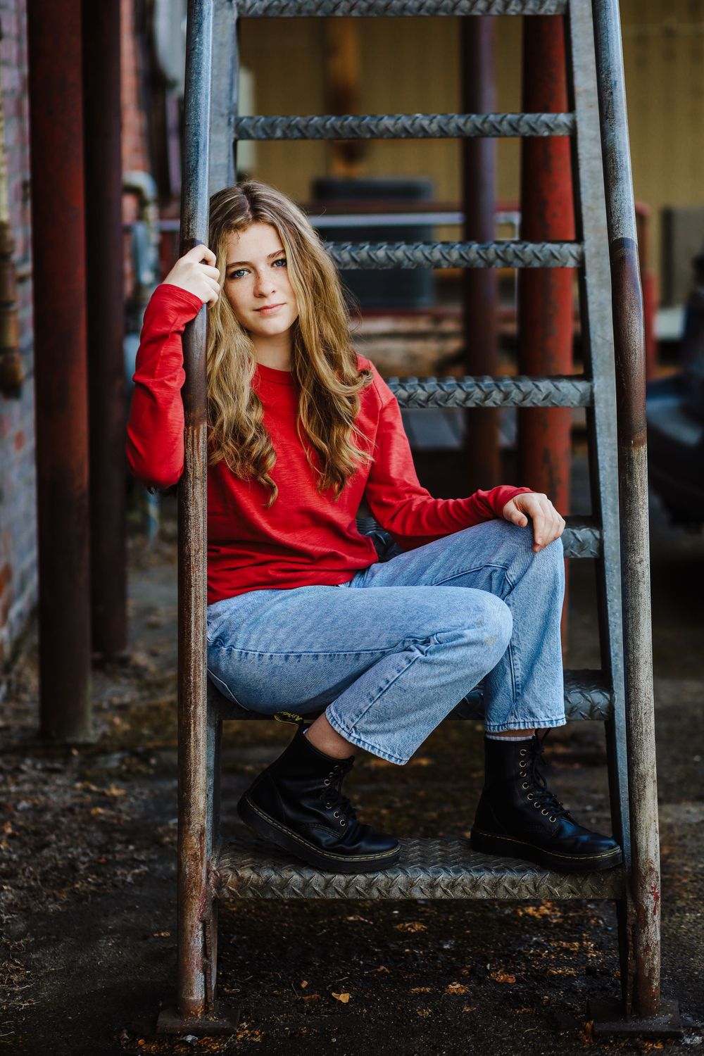 Person in red sweater and jeans sitting casually on metal ladder steps against industrial backdrop.