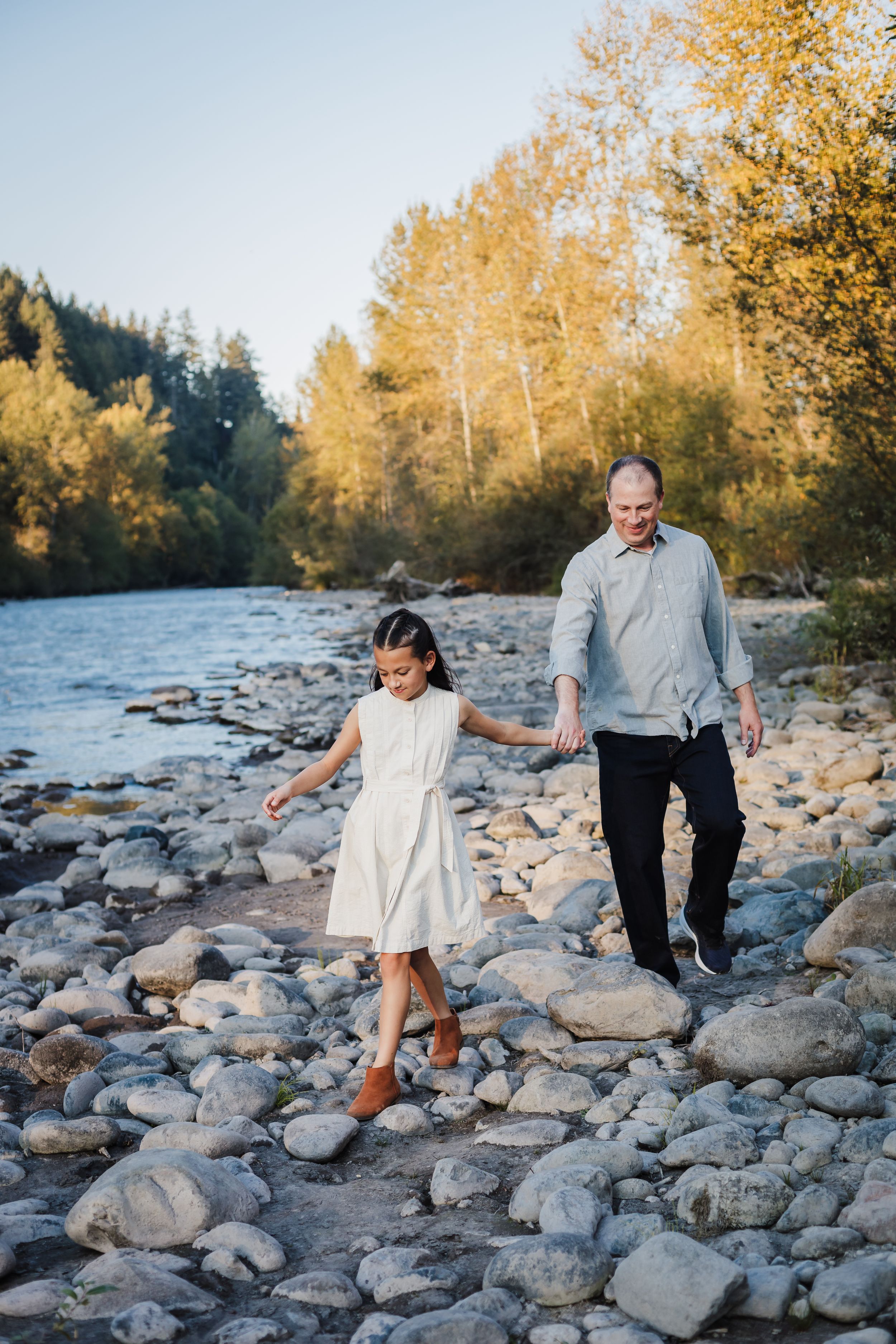 Two people walking together on rocky riverbank surrounded by golden autumn trees and flowing water.