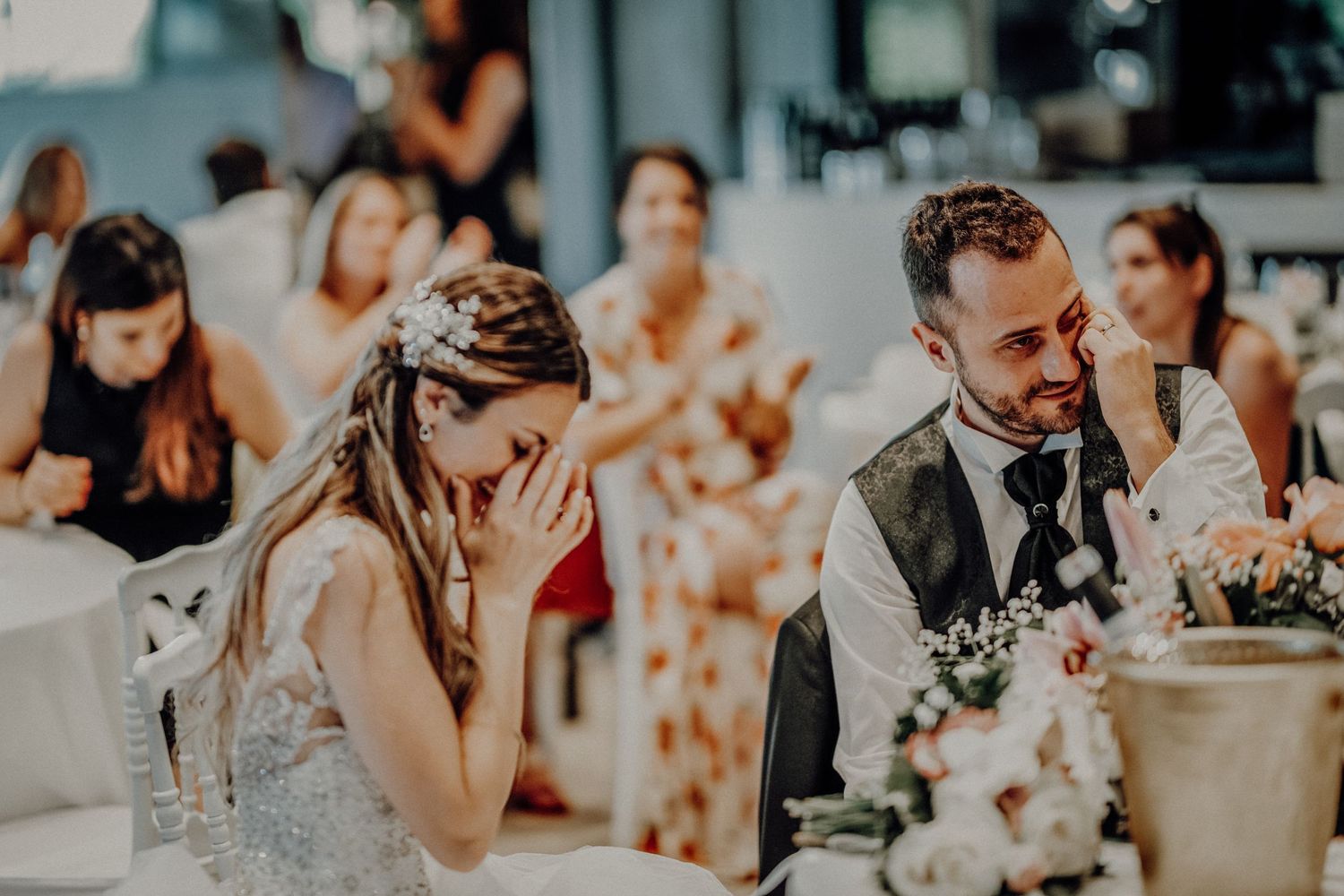 Emotional bride covering her face while groom smiles during heartfelt wedding reception speech.