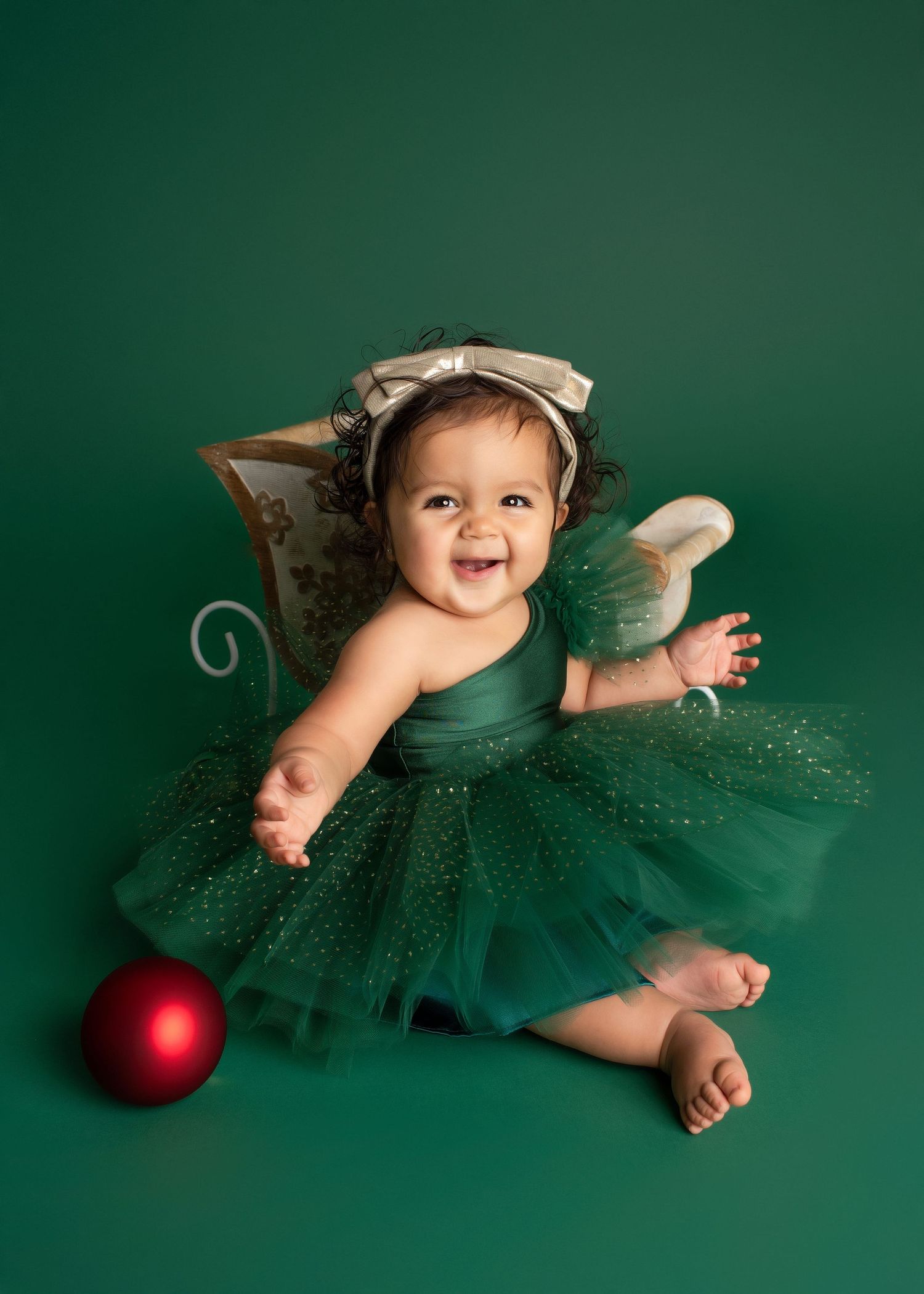 a baby smiles while wearing a green tulle dress while sitting next to a red ornament and a tiny white and gold sleigh