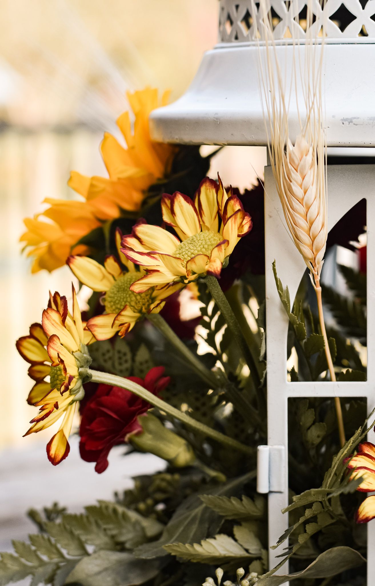 Decorative sunflowers and wheat stalks arranged next to a white lantern in soft natural lighting.