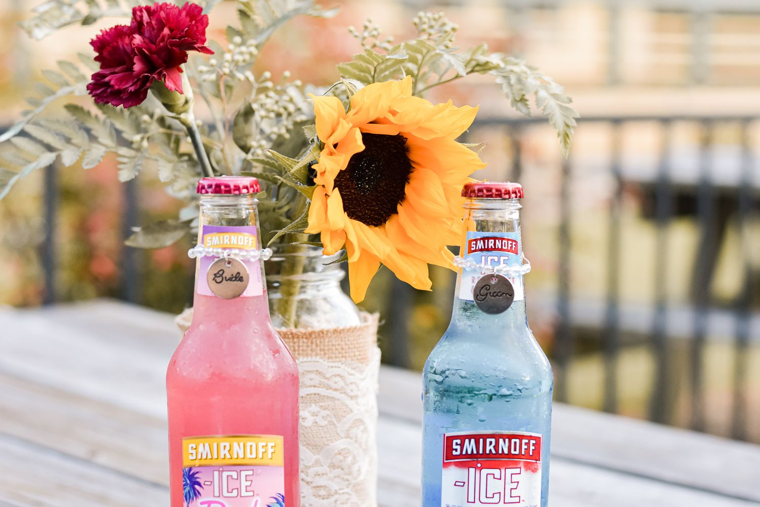 Colorful Smirnoff Ice bottles with sunflowers and carnations in a mason jar vase on a wooden deck.