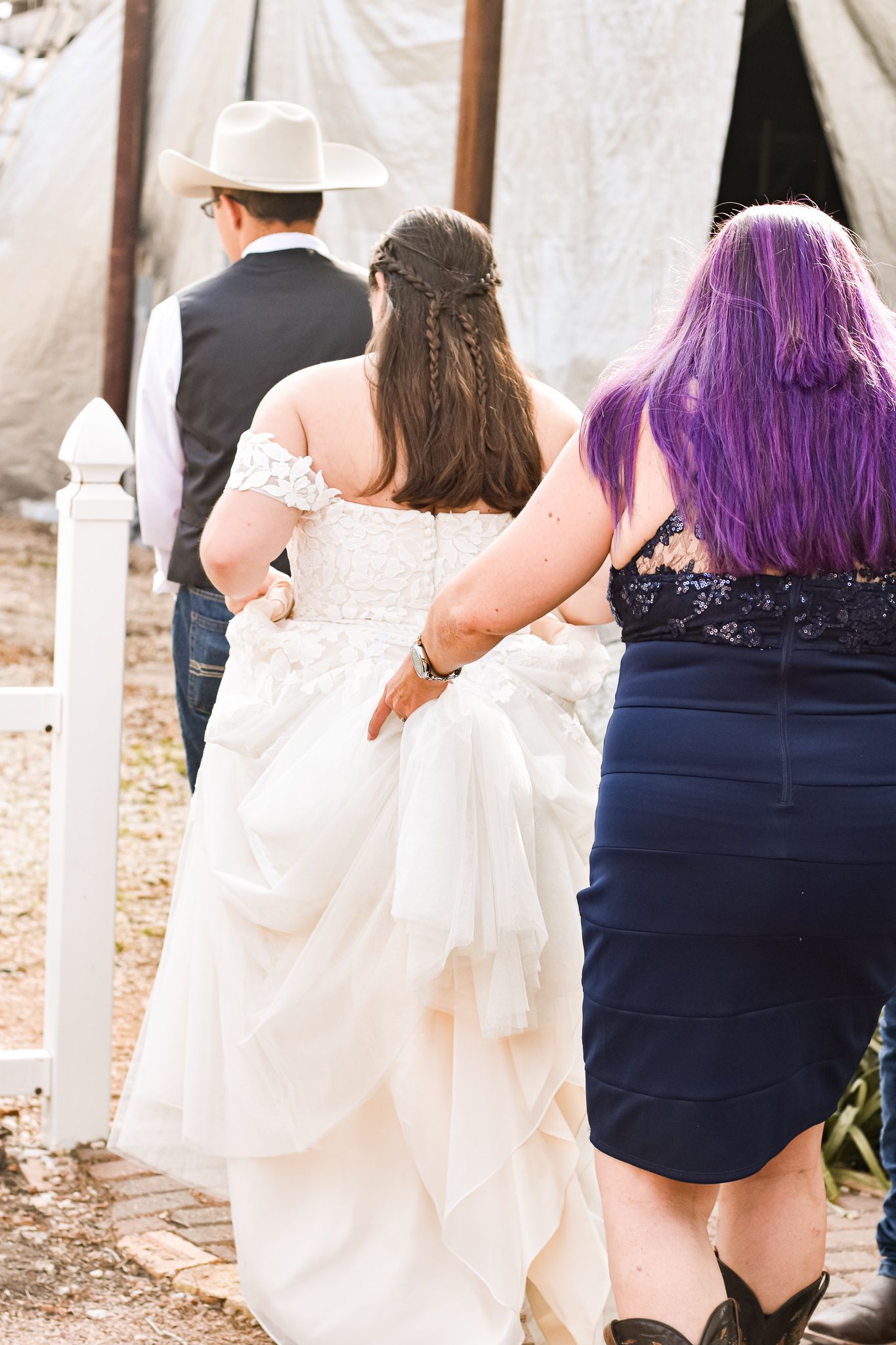 Wedding party members following a couple down a path near white tent structures.