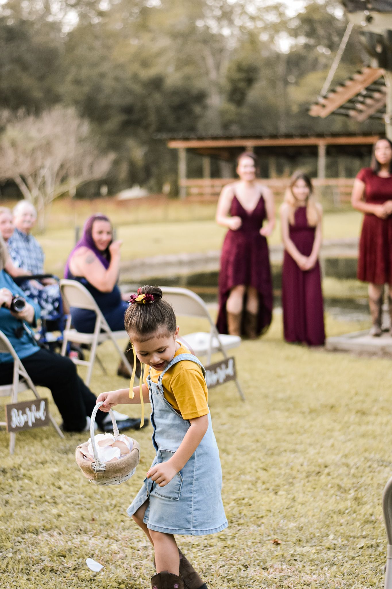 A young flower girl in yellow and denim scatters petals at an outdoor wedding ceremony.