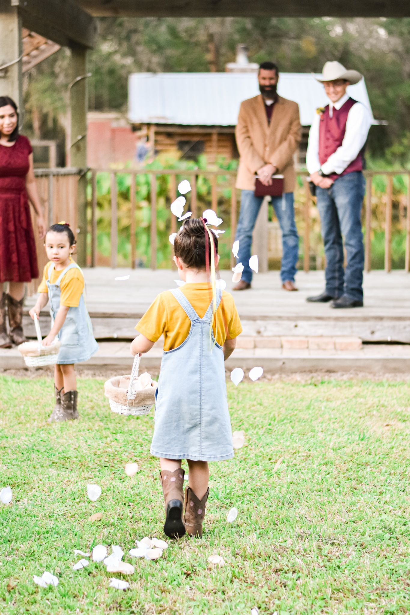 Two flower girls in matching yellow tops and denim dresses scatter petals down an outdoor aisle.