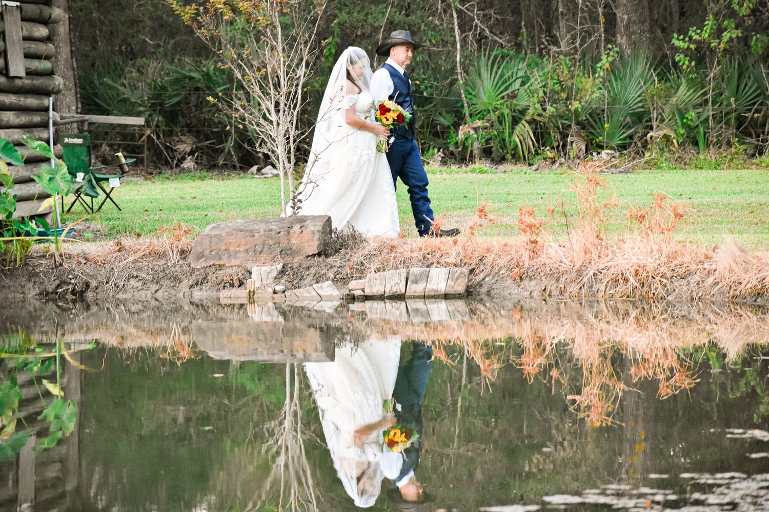 Wedding couple stands by pond creating perfect reflection in tranquil outdoor setting.