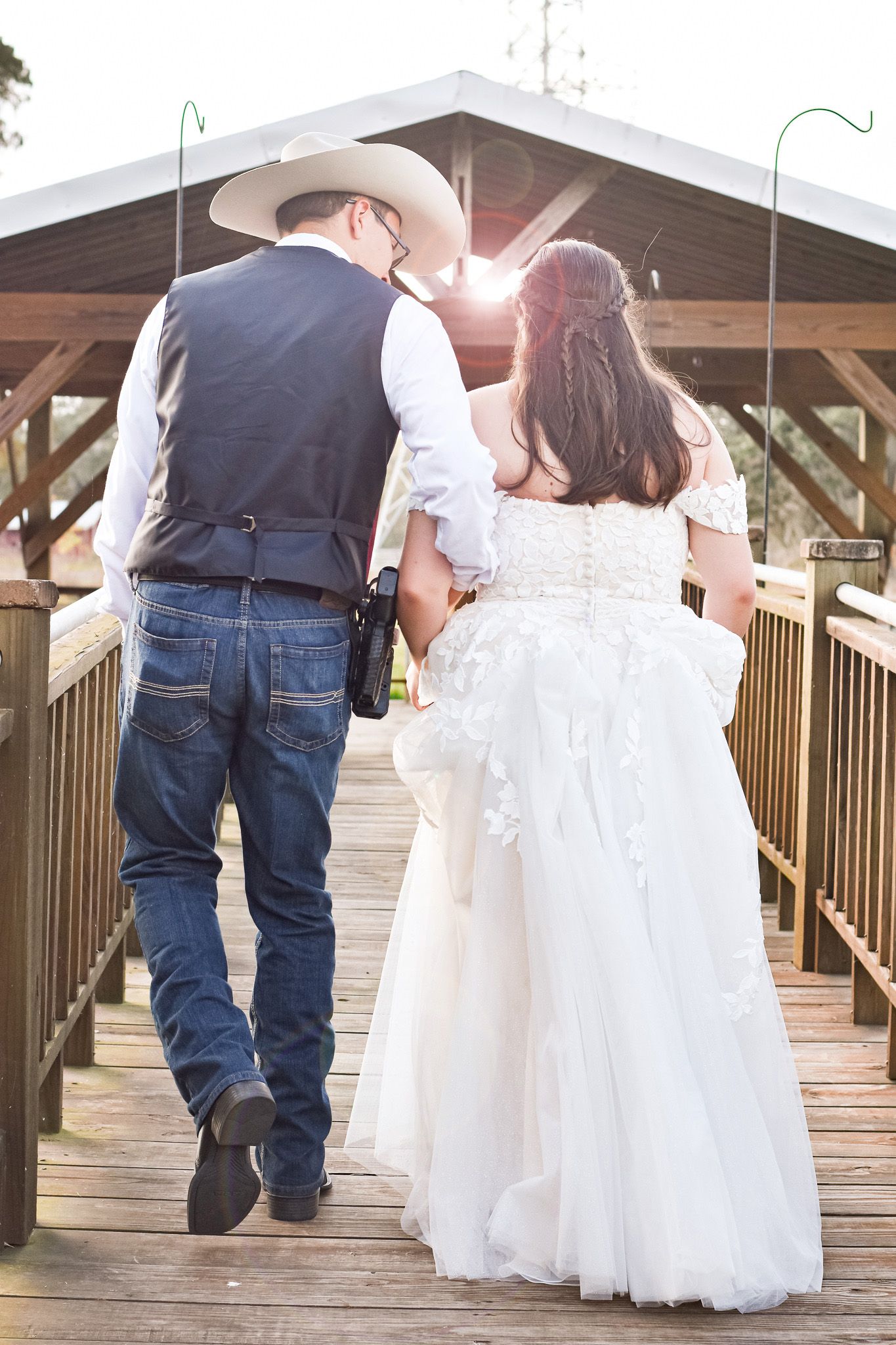 A couple in western wedding attire walking hand in hand on a wooden bridge at sunset.