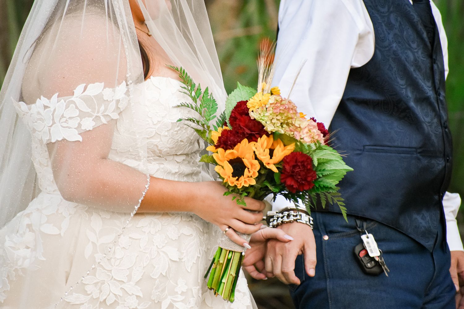 Vibrant sunflower and red rose wedding bouquet held against white lace dress and navy suit.