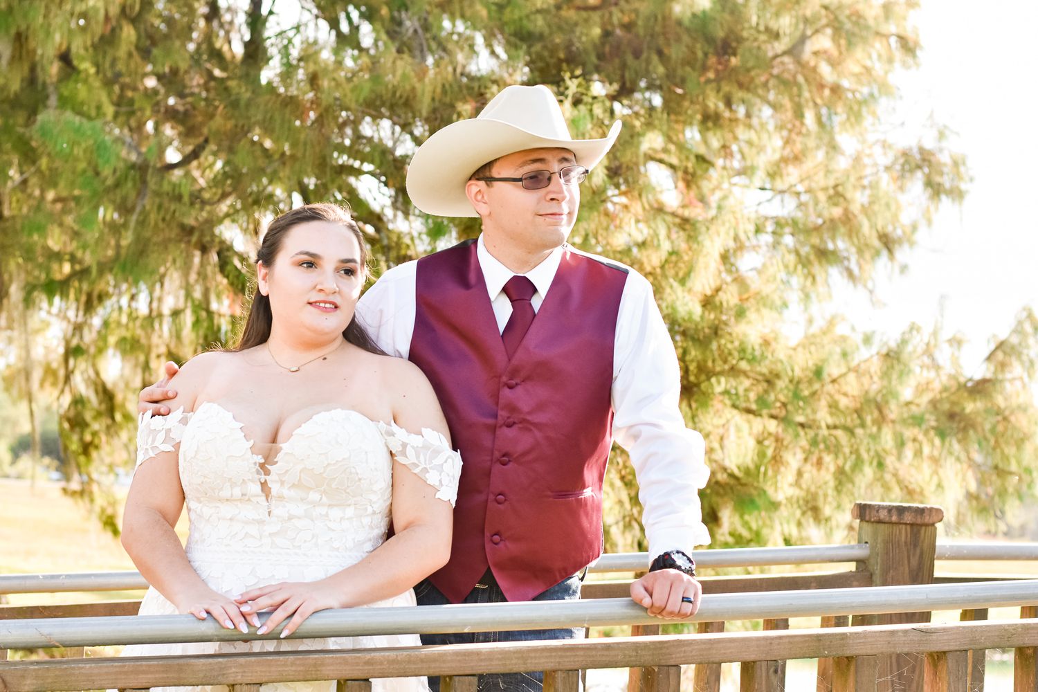 A couple in western wedding attire pose together on a wooden railing during golden hour.