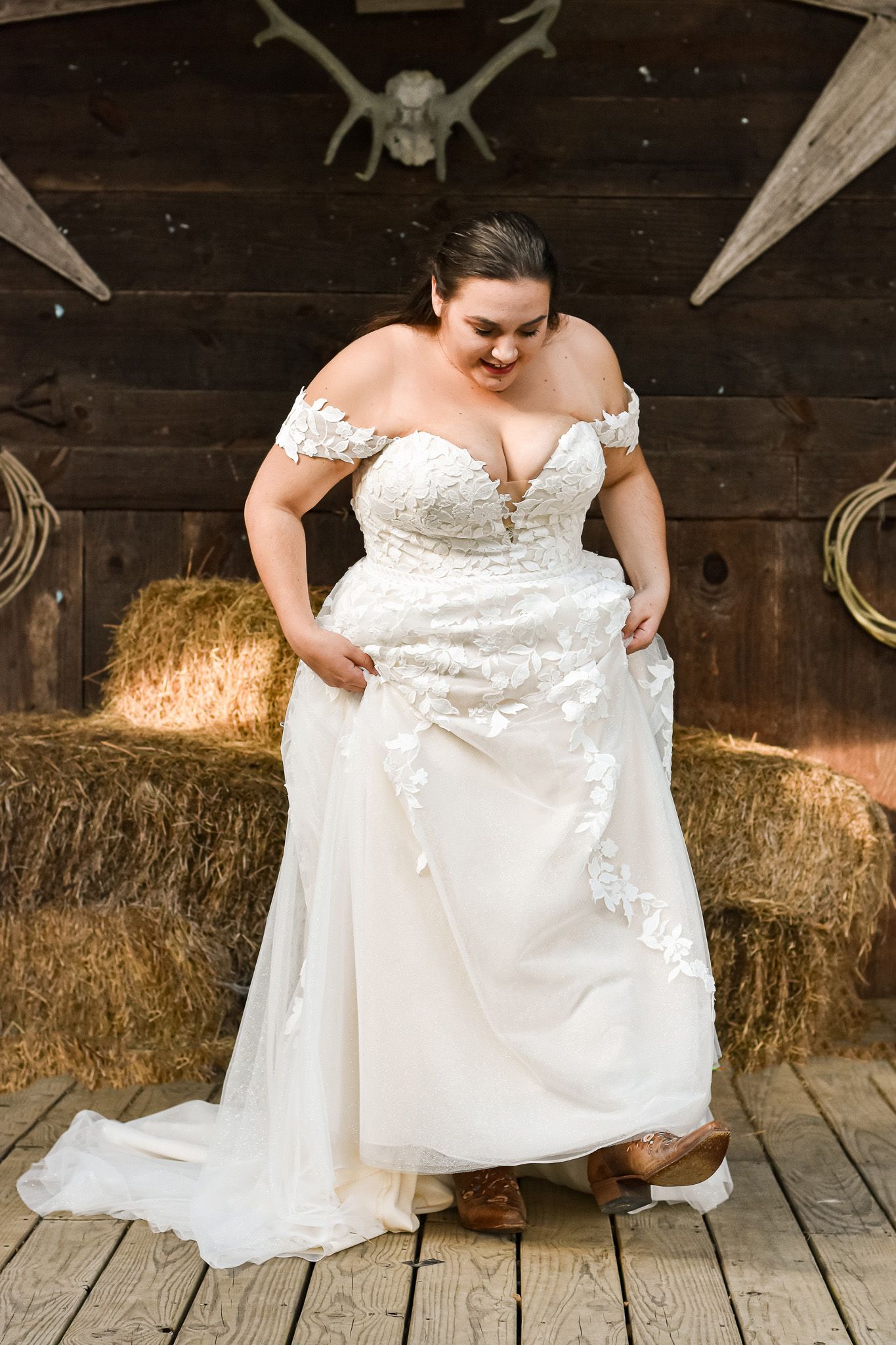 Beautiful off-shoulder lace wedding dress shown in rustic barn setting with hay bales and wooden walls.